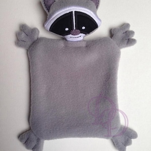 Puede incluir: Una manta de seguridad de felpa gris en forma de mapache con rasgos faciales bordados. La manta tiene cuatro brazos pequeños y dos pies pequeños.