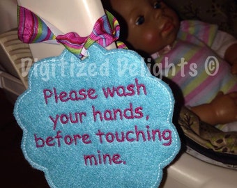 May include: A blue felt tag with the text "Please wash your hands, before touching mine." in pink embroidery. The tag is attached to a baby doll car seat.
