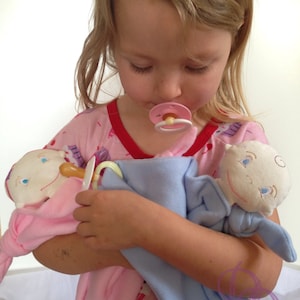 Puede incluir: Una niña pequeña con un pijama rosa y blanco sostiene dos muñecas de peluche. Una muñeca es rosa y la otra es azul. Ambas muñecas tienen chupetes en la boca.