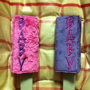 May include: Two plush fabric straps with the word "BABY" embroidered in purple on a pink strap and in pink on a purple strap.