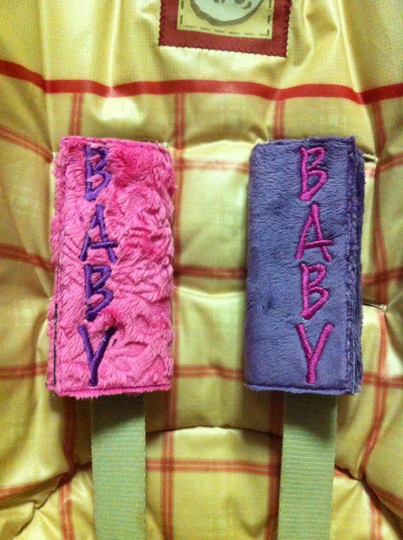 May include: Two plush fabric straps with the word "BABY" embroidered in purple on a pink strap and in pink on a purple strap.