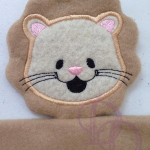 Puede incluir: Una aplicación de fieltro marrón en forma de león con una cara blanca, nariz rosa y ojos negros. El león está sonriendo con la boca abierta.
