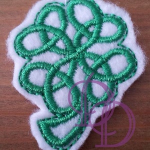 Può includere: Un design a forma di trifoglio ricamato in verde su uno sfondo di feltro bianco.