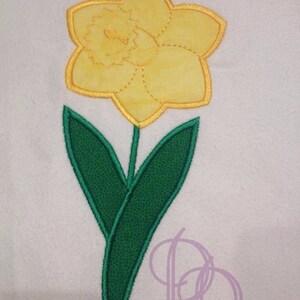 Può includere: Un narciso giallo con foglie verdi. Il fiore è ricamato su un tessuto bianco.
