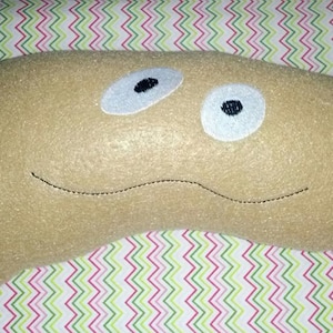 Może przedstawiać: Beżowa, pluszowa zabawka w kształcie fasoli z haftowanymi czarnymi oczami i wyszytym uśmiechem. Zabawka jest umieszczona na kolorowym, wzorzystym tle. Ta fantazyjna pluszowa zabawka ma przynieść radość.