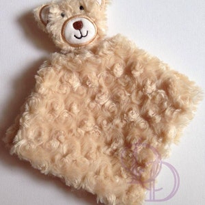 Puede incluir: Una manta de bebé de felpa beige con una cara de oso bordada en marrón en la parte superior.