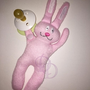 Puede incluir: Juguete de peluche rosa en forma de conejo con ojos bordados y una sonrisa cosida. El conejo tiene orejas largas y un chupete sujeto a su mano.