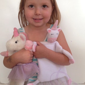 Puede incluir: Una niña pequeña sostiene dos peluches de unicornio. Uno es un unicornio blanco con detalles rosas y un tutú morado. El otro es un unicornio blanco con detalles rosas y una manta blanca.