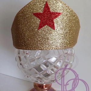 May include: A gold glitter crown with a red star emblem. The crown is set atop a clear, diamond-cut glass bowl with a rose gold base. The crown is likely for a costume or party.