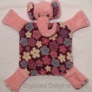 Puede incluir: Manta de bebé con estampado floral rosa y morado con una cabeza de elefante rosa unida. El elefante tiene una pequeña trompa rosa y orejas grandes. El texto "Digitized Delights" es visible en la parte inferior de la imagen.