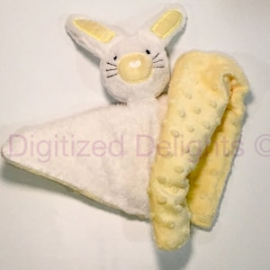Puede incluir: Una manta de seguridad de conejito blanco y amarillo con una tela amarilla texturizada. El conejito tiene ojos, nariz y boca bordados.