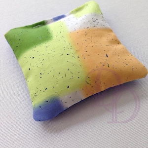 May include: A small, square-shaped pillow with a colorful, tie-dye pattern. The pillow is made of fabric with a speckled texture and features shades of green, blue, orange, and white.