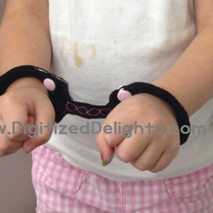 May include: A pair of black fabric handcuffs with pink buttons. The handcuffs are connected by a black strap with pink stitching.