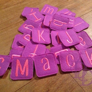 May include: A set of purple felt squares with pink letters embroidered on them. The letters spell out the word "Mack" and include other letters of the alphabet.