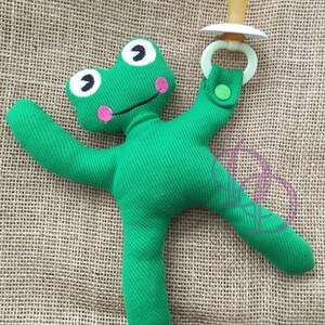 Puede incluir: Juguete de peluche verde en forma de rana con un chupete unido a un brazo. El juguete tiene ojos bordados y una boca sonriente.