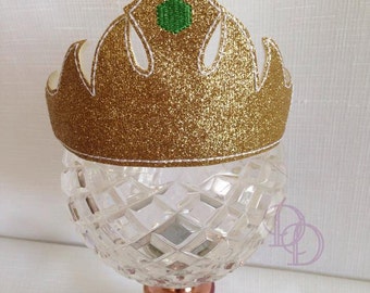 May include: A gold glitter crown with a white outline and a green gem in the center. The crown is sitting on a clear glass pedestal with a copper base.