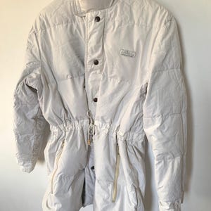 May include: A cream-colored puffer jacket featuring a high collar, a front button closure, and a drawstring waist. The jacket includes two vertical zippered pockets on the front and a small embroidered logo on the upper chest area.