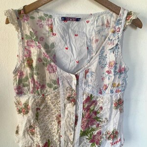 Peut inclure: Un haut sans manches boutonné avec un motif patchwork composé de divers imprimés floraux roses, verts et bleus sur fond blanc. Le vêtement présente un petit logo Replay rouge sur le côté inférieur droit et des détails à volants aux épaules.