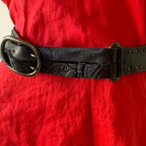 Peut inclure: Une ceinture en cuir noir avec une surface texturée et une grande boucle ovale. La ceinture a une conception à double sangle et est ornée de petits clous ronds le long des bords. La ceinture est portée sur un vêtement rouge.