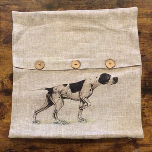 May include: A light gray linen pillow cover featuring a black and white illustration of a Pointer dog. The pillow has three wooden buttons and a flap closure. The dog is depicted in a walking pose.