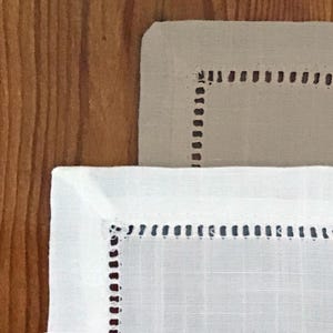 May include: Close-up of two linen napkins. One is white, the other is beige. Both have a stitched border with small, evenly spaced dark brown stitches. The napkins are on a wooden surface.