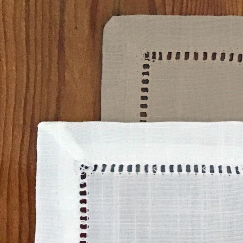 May include: Close-up of two linen napkins. One is white, the other is beige. Both have a stitched border with small, evenly spaced dark brown stitches. The napkins are on a wooden surface.