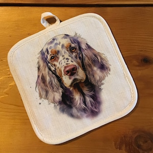May include: A white cotton pot holder with a brown and white spotted dog with brown eyes. The dog is looking directly at the camera.