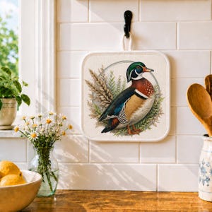 May include: Square, off-white pot holder with a colorful illustration of a wood duck. The duck is surrounded by wheat and greenery, set within a circular frame. A black hook and white loop are attached for hanging.