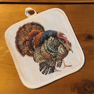 May include: Square off-white pot holder with a loop, featuring a watercolor illustration of a turkey. The turkey has brown, red, and blue feathers. The pot holder has a white border and is on a wooden surface.