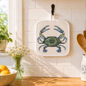 May include: Square off-white pot holder with a blue crab design. The crab is depicted in shades of blue and green, with its claws extended. The pot holder has a white border and a loop for hanging.