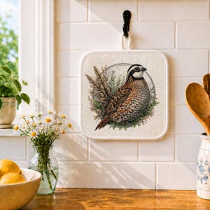 May include: Square beige pot holder with a detailed illustration of a quail bird surrounded by wheat and greenery. The bird has brown, white, and black feathers. The pot holder hangs from a black hook on a white tiled wall.