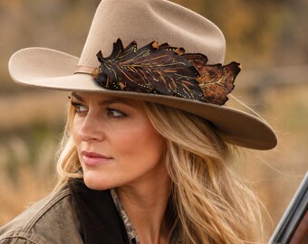 May include: A beige felt fedora hat featuring a wide brim and a decorative dark brown leather accent shaped like a large oak leaf. The leaf detail is embellished with fine gold-colored stitching along the veins and edges of the material.