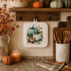 May include: A square off-white fabric potholder featuring a watercolor-style illustration of three pumpkins in shades of teal, white, and orange, surrounded by autumn leaves, hanging from a black hook on a wooden rack against a white wall.