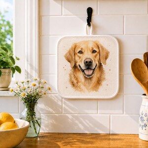 May include: Square beige pot holder featuring a watercolor portrait of a golden retriever dog. The dog has a happy expression with its tongue out. The pot holder has a white border and a loop for hanging. A decorative kitchen item.
