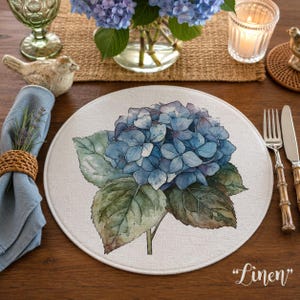 May include: A round white placemat featuring a watercolor illustration of a blue hydrangea flower with green leaves. The placemat is positioned on a wooden table next to a blue cloth napkin in a woven ring, a fork, and a knife.