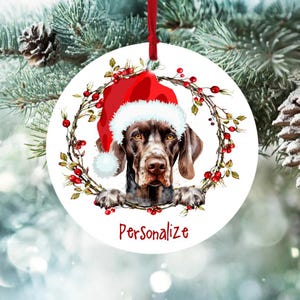 May include: Round Christmas ornament featuring a brown and white dog wearing a red Santa hat. The dog's head and paws are visible, surrounded by a wreath of berries and greenery. The word "Personalize" is written in red below the dog.