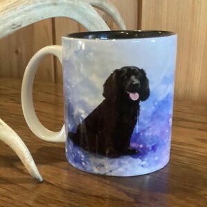 May include: White ceramic mug with a black interior and a white handle. The mug features a watercolor design with a black dog as the focal point. The background has blue and purple hues.
