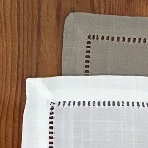 以下が含まれることがあります： 装飾的なステッチの縁取りが施されたリネン製のナプキン2枚。上のナプキンは薄いグレーで、下のナプキンは白です。