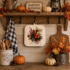 May include: A square fabric pot holder featuring a printed autumn arrangement of pinecones, red berries, and fall foliage. The pot holder hangs from a wooden hook on a rustic wall shelf decorated with pumpkins, a lantern, and a sign reading Harvest Blessings.