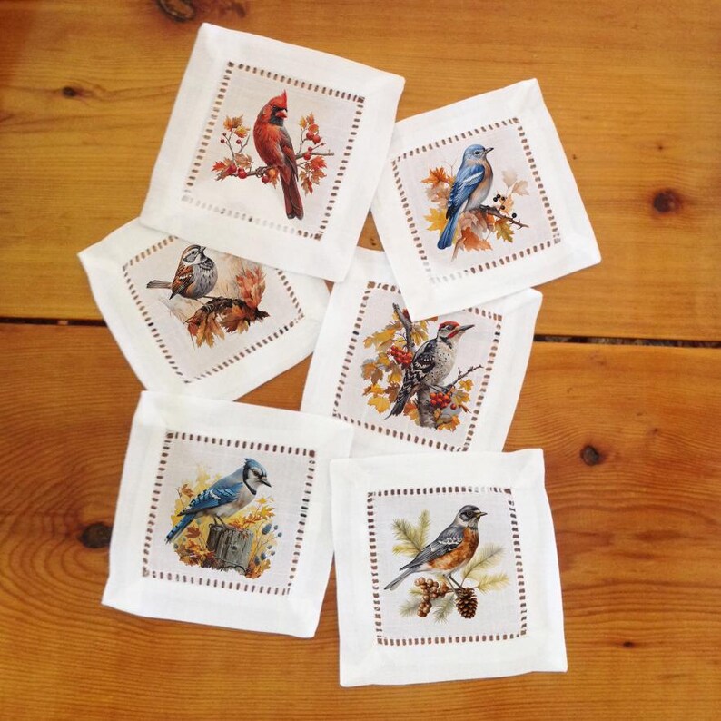 May include: A collection of six white linen coasters, each featuring a different detailed illustration of a bird. The birds include a cardinal, bluebird, woodpecker, and others, set against autumn foliage. The coasters have a stitched border.