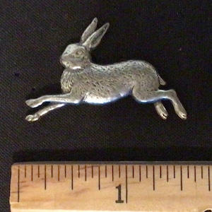 May include: A silver-colored metal hare figurine in a running pose. The hare is approximately 1 inch long, with detailed features including long ears and a textured body. The figurine is set against a dark background.