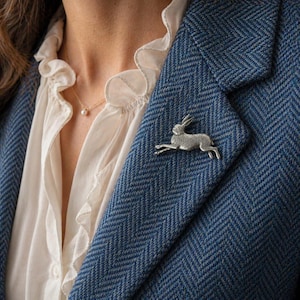 May include: A silver-colored brooch in the shape of a running hare is pinned to a blue herringbone blazer. The blazer is worn over a white blouse with a ruffled collar. A delicate gold necklace with a small pearl pendant is also visible.
