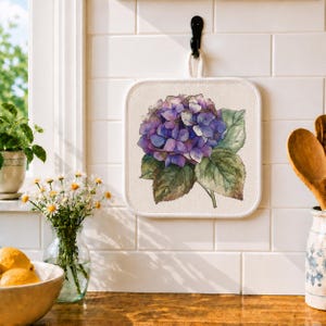 May include: Square, off-white pot holder with a watercolor painting of a purple hydrangea flower and green leaves. The pot holder has a white border and a loop for hanging. It hangs on a black hook against a white tile wall.