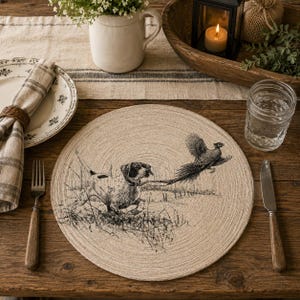 May include: A round, beige woven placemat featuring a black ink-style illustration of a hunting dog chasing a pheasant through tall grass. The placemat has a textured, circular coil pattern and is centered on a dark wooden table surface.