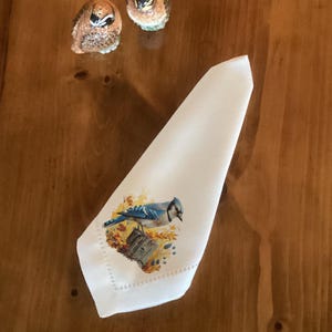 May include: White cloth napkin with a blue jay illustration perched on a tree stump surrounded by autumn leaves. Two owl-shaped salt and pepper shakers are in the background. The napkin has a decorative stitched edge.