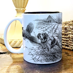 May include: White ceramic coffee mug with a black rim and handle. The mug features a black and white illustration of a dog pointing at a pheasant in a field.