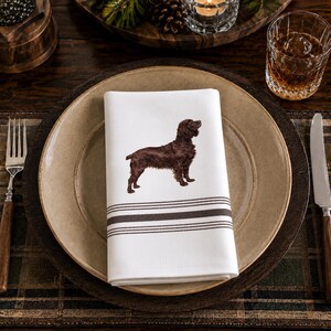 May include: A white cloth napkin featuring a brown dog illustration in the center and three horizontal dark brown stripes near the bottom edge. The napkin is folded neatly and placed on a round, tan-colored ceramic dinner plate.