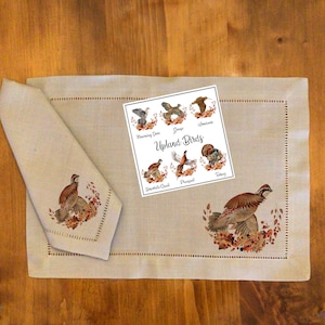 以下が含まれることがあります： ベージュのリネン製のプレースマットに、茶色と白の刺繍で鳥のデザインが施されています。プレースマットの上に、「Upland Birds」という文字と、さまざまな鳥のイラストが描かれたカードが置かれています。