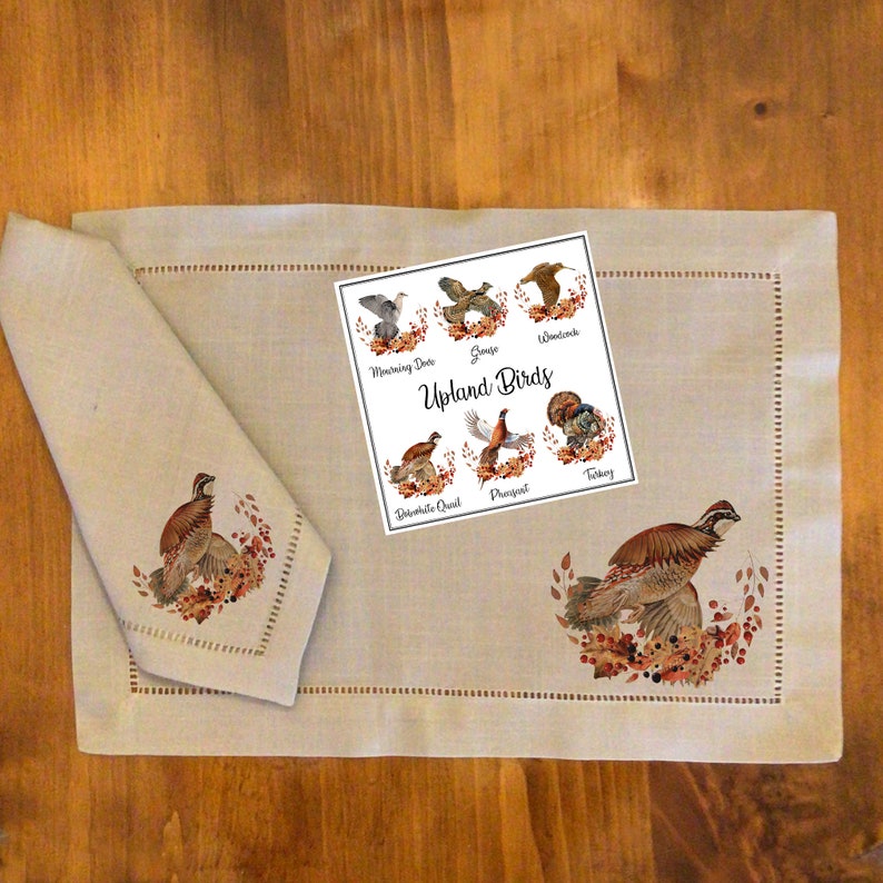以下が含まれることがあります： ベージュのリネン製のプレースマットに、茶色と白の刺繍で鳥のデザインが施されています。プレースマットの上に、「Upland Birds」という文字と、さまざまな鳥のイラストが描かれたカードが置かれています。