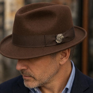 May include: A brown fedora hat with a matching band and a decorative dog-shaped pin. The hat has a classic brim and a structured crown. The hat is made of a felt-like material.